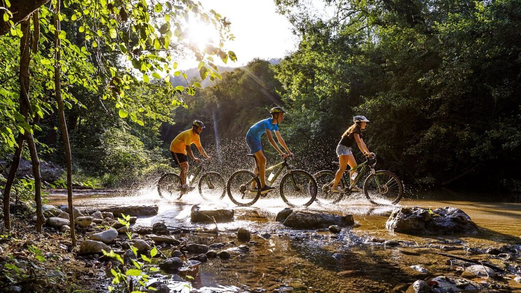 Drei Jugendliche fahren in Spanien mit dem Mountainbike durch einen flachen Fluss im Ferienlager