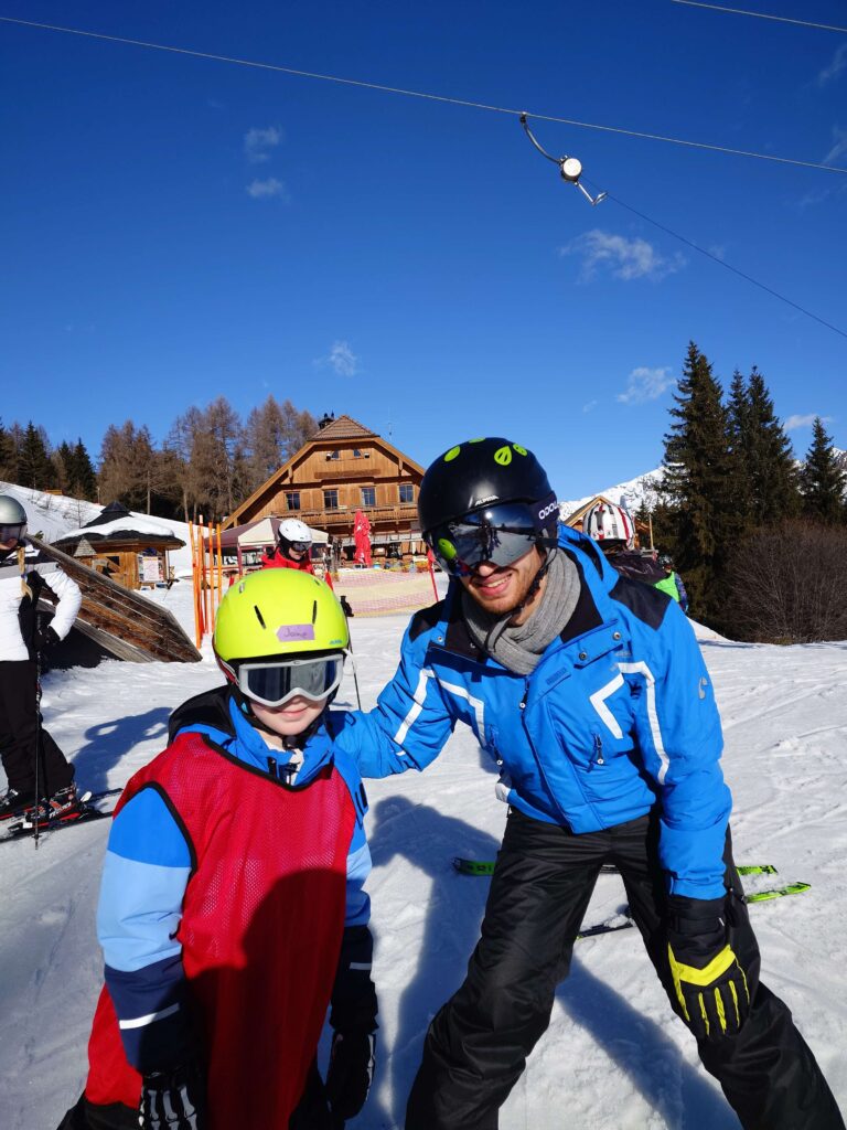 Kind mit gelbem Helm und Betreuer*in im Schnee vor Skihütte beim Wildfang e.V. Wintercamp – betreute Ferienlager-Jugendreise mit Skiausflug