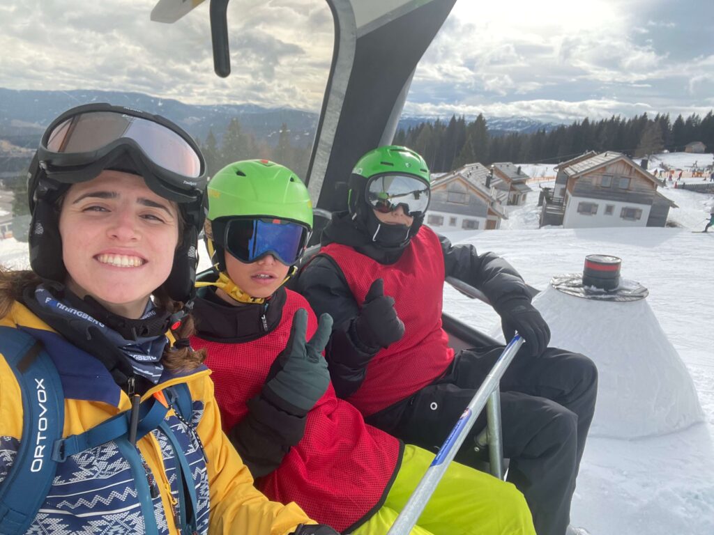 Jugendbetreuer*in mit zwei Kindern im Skifreizeit-Ferienlager von Wildfang e.V. auf dem Sessellift, lachende Gesichter, Skihelm und Winterlandschaft – Ferienlager Betreuer Alltag, pädagogische Jugendreise und Betreuung im Schneesportcamp