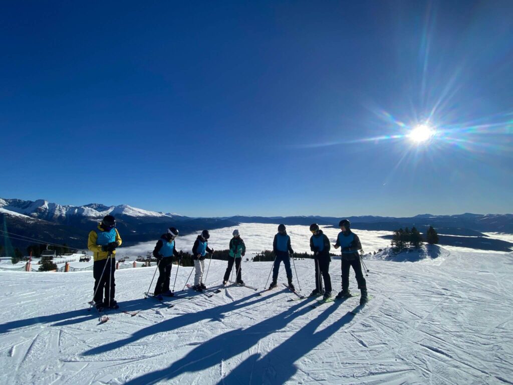 Kinder und Jugendliche im Skiurlaub-Ferienlager von Wildfang e.V. stehen als Gruppe auf der Skipiste mit Betreuer*in im Wintercamp, klare Berglandschaft und blauer Himmel – Ferienlager Betreuer Alltag, Winterferienreise und Jugendreise mit Betreuung