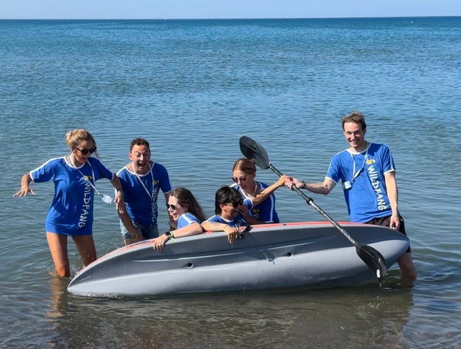 Sechs Teamer in blauen Wildfang-T-Shirts stehen lachend mit einem Surfbrett im Meer